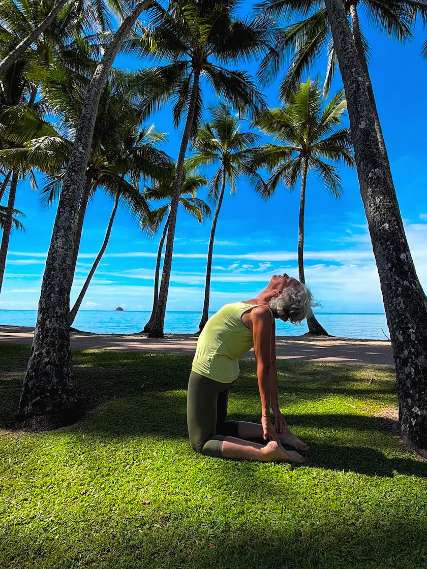 Susan Stackhouse kneeling in camel pose, Ustrasana, on the grass under the palms at Kewarra Beach, head back and hands on her heels