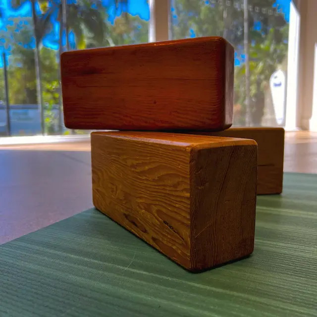 Two timber yoga blocks stacked on a green mat, with palms outside the studio window behind them