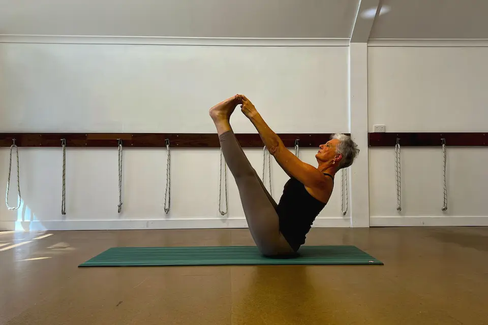 Susan Stackhouse seated on a mat in the studio, holding both feet with her legs raised in Ubhaya Padangusthasana