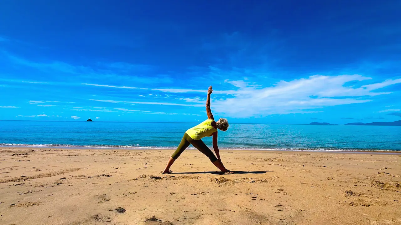 Susan Stackhouse in extended triangle pose, Utthita Trikonasana, on the sand at Kewarra Beach with the sea and sky behind her